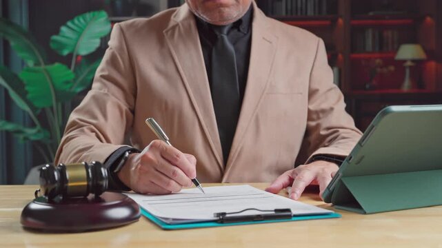 Mature lawyer sitting inside modern office writes notes over paper clipboard beside wooden gavel digital tablet reviewing judicial contract legal case.