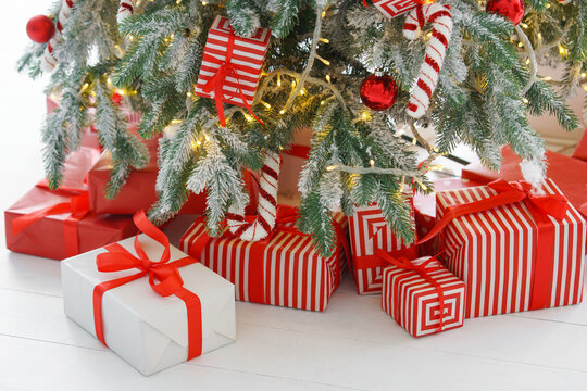 Beautiful presents, gift boxes wrapped in elegant red paper under green snowy festive Christmas tree with warm lights. Cozy winter holiday atmosphere perfect for seasonal celebrations and decor themes