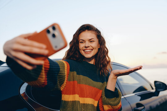Smiling woman taking selfie with smartphone near car at sunset. Concept of modern lifestyle, travel, and mobile communication. Bright emotions and happy mood create a warm atmosphere outdoors.