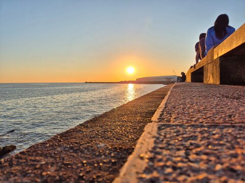 People sitting along the Rambla seawall while watching a golden sunset over the Rio de la Plata in Montevideo, Uruguay.