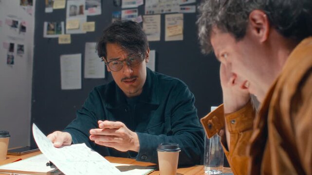 Focused male filmmaker in glasses explaining script details to colleague and having discussion with team during preproduction meeting at desk in creative office