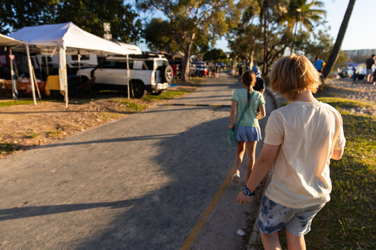 Two young people walk along a sunlit path near a beachside