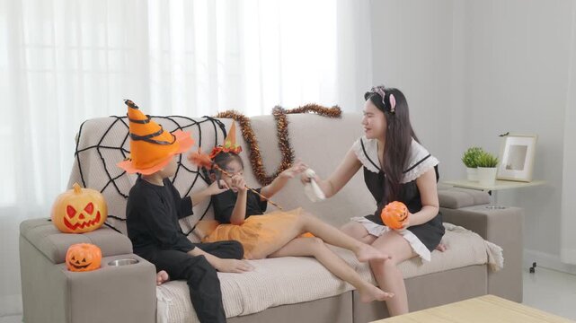 Asian family enjoys Halloween celebration at home, mother and children wear spooky costumes, holding pumpkin baskets, sitting on sofa with spiderweb decor, happy festive fun.