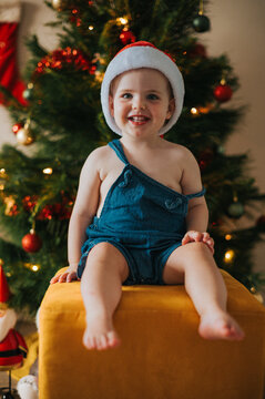 Happy blonde toddler sits on a yellow stool with a joyous grin by Christmas decorations
