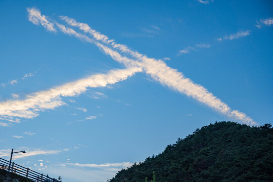Crossing white airplane contrails stretch across the clear blue sky above a dark green mountain ridge in the evening.