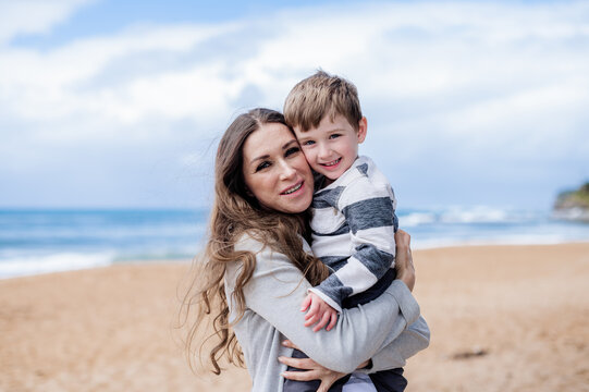 Woman embraces her smiling son at the beach with waves and sand in the background