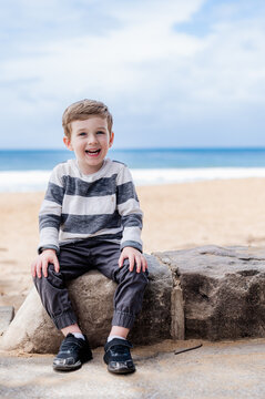 Young boy smiles while sitting on a rock by the beach, with the ocean behind him