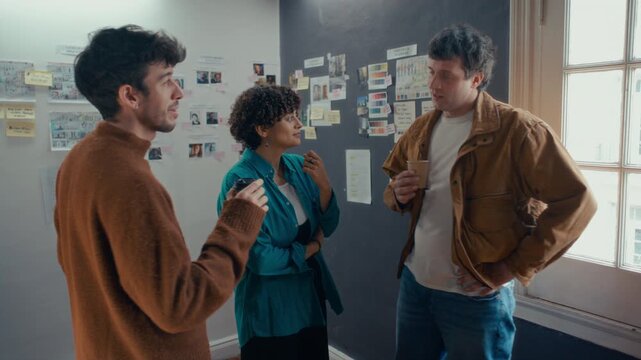 Group of three professional filmmakers drinking coffee and discussing ideas during break in creative studio with reference moodboard on walls