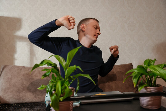 Adult man stretches arms after working at laptop in home workspace. Relaxed expression notebook pen and indoor plants create balanced work routine scene. Concept of comfortable home setting illustrate