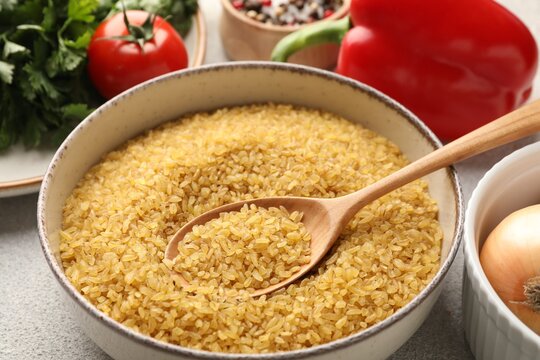 Raw bulgur in bowl, spoon and other products on table, closeup
