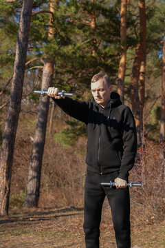Young adult man performs strength training with adjustable metal dumbbells resting on shoulders during outdoor workout in pine forest. Black sportswear, standing pose, healthy active lifestyle fitness