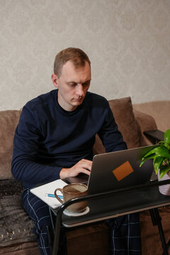 Adult man writes in notebook while looking at laptop in home workspace. Calm concentrated expression accompanies organized desk with computer pen and coffee mug. Concept of remote work, online