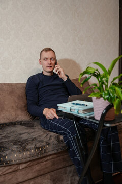 Adult man talks on smartphone while writing notes beside laptop at small table in home interior. Focused expression, notebook pen and coffee mug create productive remote work scene. Warm indoor light