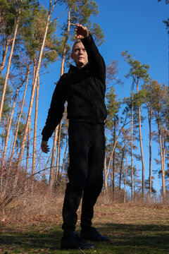 Young adult man performs standing side stretch during outdoor exercise in pine forest. Black athletic clothing, full body view flexibility training, warmup mobility, healthy active lifestyle copy