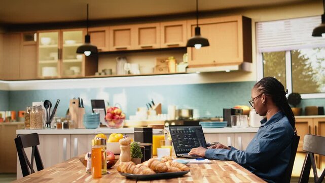 Black woman programs on laptop in kitchen during hybrid IT work, writing source code with command-line tools and cloud computing for software development.