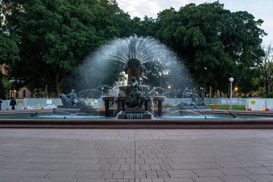 Water spraying from public park fountain featuring bronze statues surrounded by lush green trees