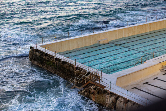 Empty coastal ocean bath pool filled with calm blue water beside crashing rough sea waves