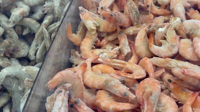 Close-up supermarket freezer display with transparent divider. Raw grey shrimps on left and cooked pink prawns on right covered in ice crystals. Grocery store seafood selection, healthy frozen food.