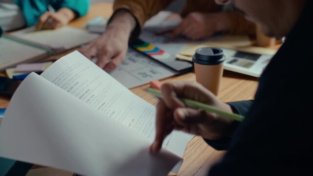 Filmmaker using pencil to mark up printed script pages during preproduction meeting with creative team at office table. Close up view