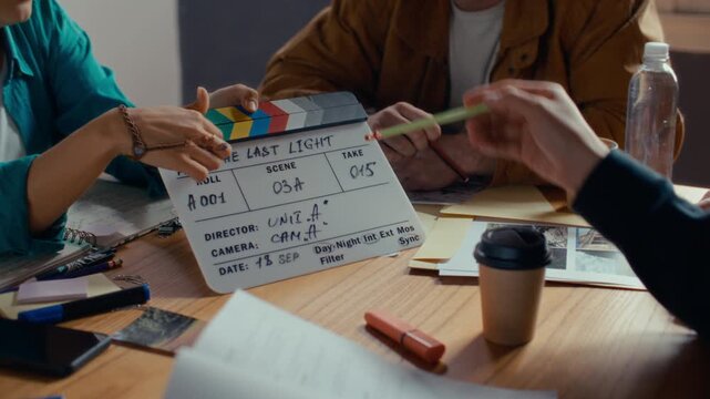 Hands of film production team members reviewing information on clapperboard and printed location photos on desk during preproduction meeting. Close up shot