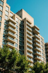 Obraz premium Modern apartment block with concrete balconies, glass railings. High rise residential building stands under clear blue sky. Green trees grow in front of urban housing structure. Contemporary city
