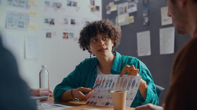 Female costume designer holding up printed costume sketch and color swatches, presenting options to her film crew during preproduction meeting