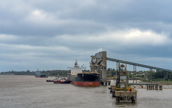 Large cargo ship docked at an industrial port terminal receiving bulk cargo loading operations via conveyor structure under overcast sky, with tugboats assisting in brown river water