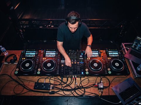 DJ at Work: A focused DJ in headphones operates a complex sound system, skillfully mixing music for a lively audience in a dimly lit club, creating an electrifying atmosphere.