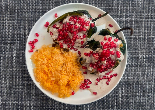 A beautifully plated serving of two Chiles en Nogada, featuring a walnut cream sauce and vibrant pomegranate seeds, accompanied by a side of Mexican red rice.