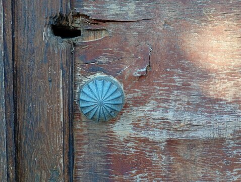 A close up of a vintage round metal door knob with a textured ribbed starburst pattern, attached to an old weathered wooden door featuring deep cracks, scratches, and a broken keyhole slot.