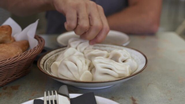 Khinkali. Man picks up a khinkali dumpling from a plate, preparing to eat it, with a basket of bread and a simple table setting visible