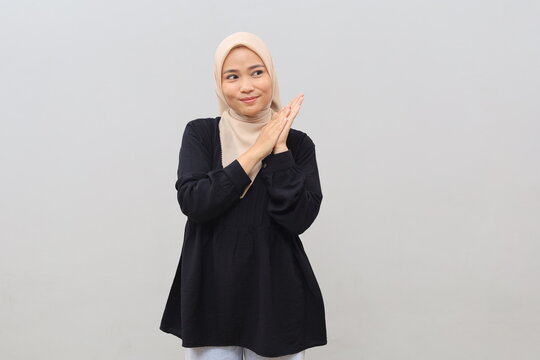 A young Asian woman wearing a headscarf and looking shrewd rubs her hands together, plotting something with a cunning expression while glancing toward an empty space with a smile.