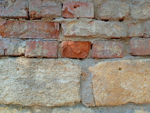 A detailed texture of an old weathered wall combining layers of destroyed red brick masonry and large porous yellow limestone blocks with thick grey cement morta