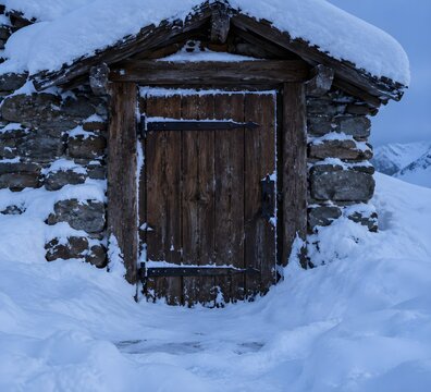 Mountain hut door drifted shut by snow with heavy timber planks and iron hinges buried to the lintel