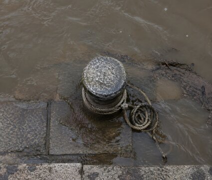 Weathered stone bollard rising from the submerged deck of a flooded quay with a slack rope coil