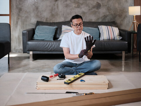 One Asian mature man wearing protective gloves prepares DIY tools and flat-pack furniture for assembly, builds for home improvement, wooden craft install hobby workshop, handyman active lifestyle.