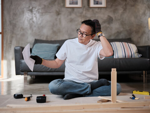 Asian mature man self-assembling flat-pack furniture with DIY tools and instruction guide, confused with wooden parts, home improvement, craft installation, hobby workshop, handyman active lifestyle.