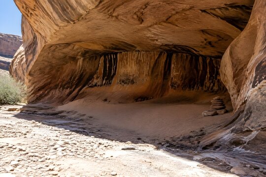 Deep sandstone alcove over a dry arroyo with a swept floor of fine sand and stacked flat stones at the drip line