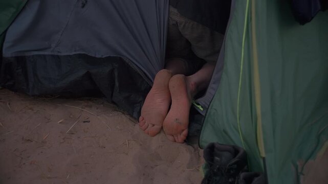 Bare feet stick out from beneath a tent flap onto the sand, capturing a relaxed moment of rest during a beach camping trip.