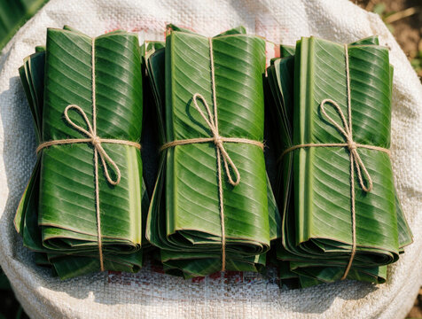 Bundles of fresh green banana leaves tied with rough twine sit on a textured white sack. They are neatly stacked, ready for use in cooking or packaging food.