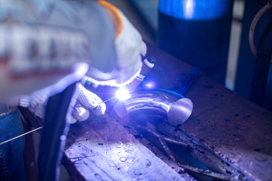 Selective focus to hands of professional welder using argon TIG process in a dark environment illuminated by the bright welding arc, welding a stainless steel curved pipe elbow on a workbench.