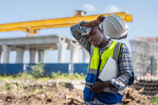 Tired construction worker man in hard hat and safety vest wiping sweat at building site, exhausted engineer holding laptop and walkie talkie taking off helmet during hot day at construction yard