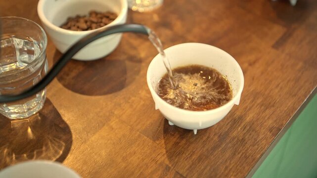A barista is pouring hot water into a cup containing finely ground coffee beans, preparing it for a Q-Grader to taste