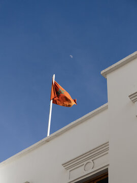 Moroccan national flag waving on a white building with the daytime moon in a clear blue sky