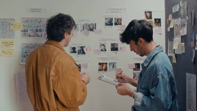 Two creative filmmakers analyzing casting photos on wall moodboard and taking notes on clipboard during preproduction work in studio. Back view