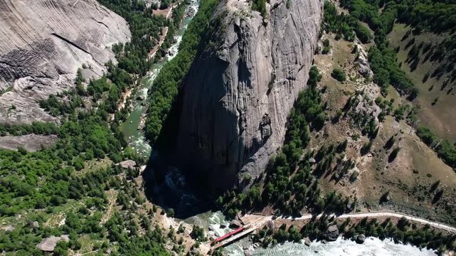 Aerial drone view of the iconic Shen Zhong Shan rock formation in Keketuohai Scenic Area, Xinjiang, with turquoise river winding through forested valley and red tourist train on bridge
