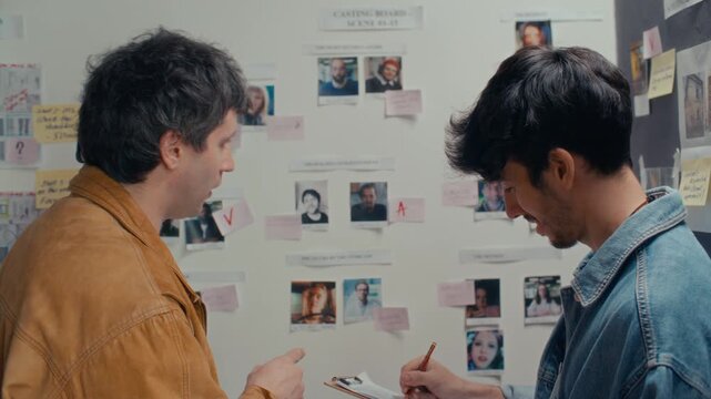 Two young creative filmmakers discussing character roles at casting board wall and writing down notes during film preproduction session in studio. Rear view