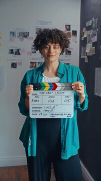 Young female filmmaker with curly hair holding clapperboard and posing for camera with smile in front of preproduction moodboard wall in creative studio. Vertical portrait