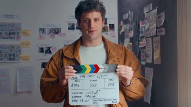 Young male filmmaker holding movie slate and posing for camera in front of reference moodboards on wall in office. Video portrait
