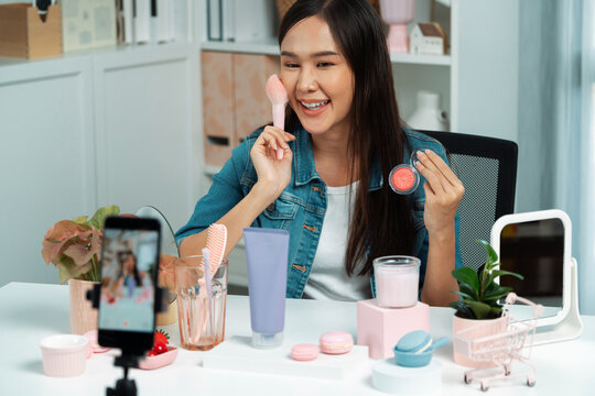 Young beautiful Asian reviewing cosmetic makeup brush cheek testing collection with pastel color, showing glow blusher mood with pink to promote on social media online recording smartphone. Stratagem.